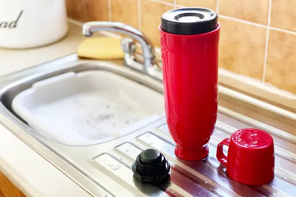 a-red-travel-mug-dismantled-for-cleaning-next-to-a-kitchen-sink-filled-with-soapy-water. a-red-travel-mug-dismantled-for-cleaning-next-to-a-kitchen-sink-filled-with-soapy-water.